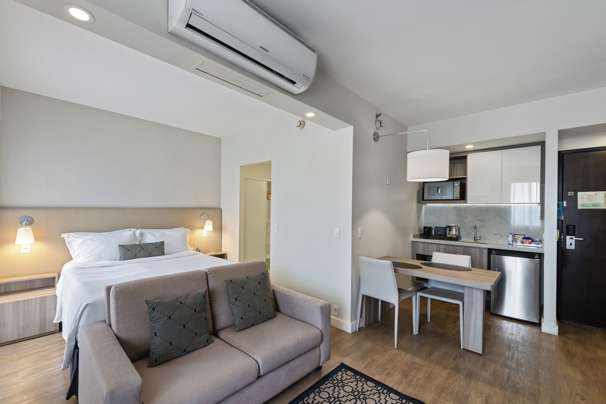 A modern hotel room with a bed, sofa, desk, and kitchenette.