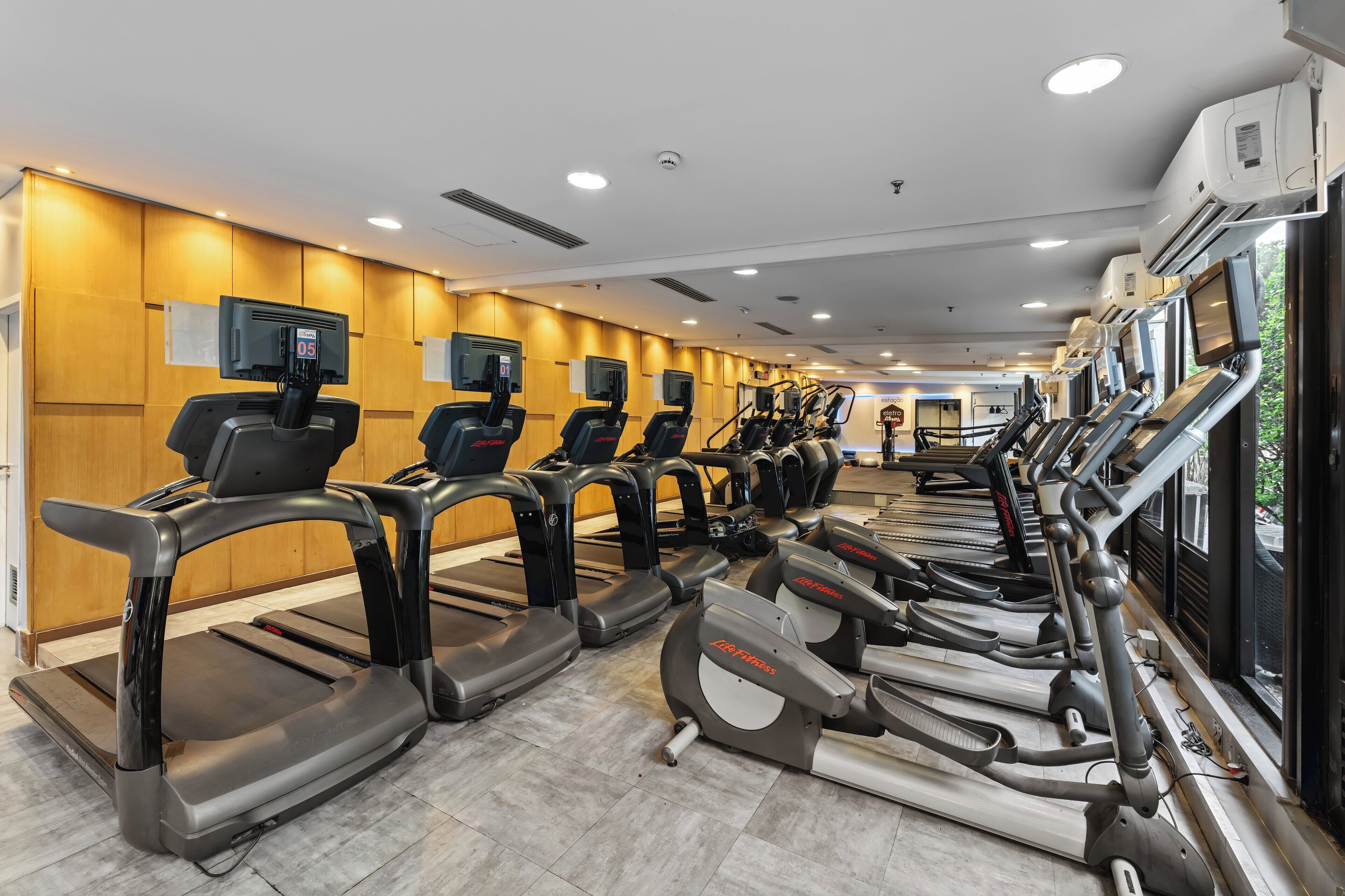 Une salle de sport équipée de plusieurs tapis de course et de vélos d’exercice, avec des murs recouverts de panneaux en bois et des unités de climatisation fixées au plafond.