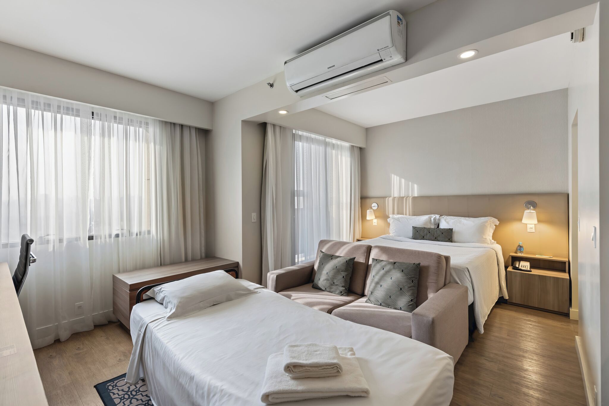 A hotel room with two beds, a sofa, a desk, and an air conditioning unit.