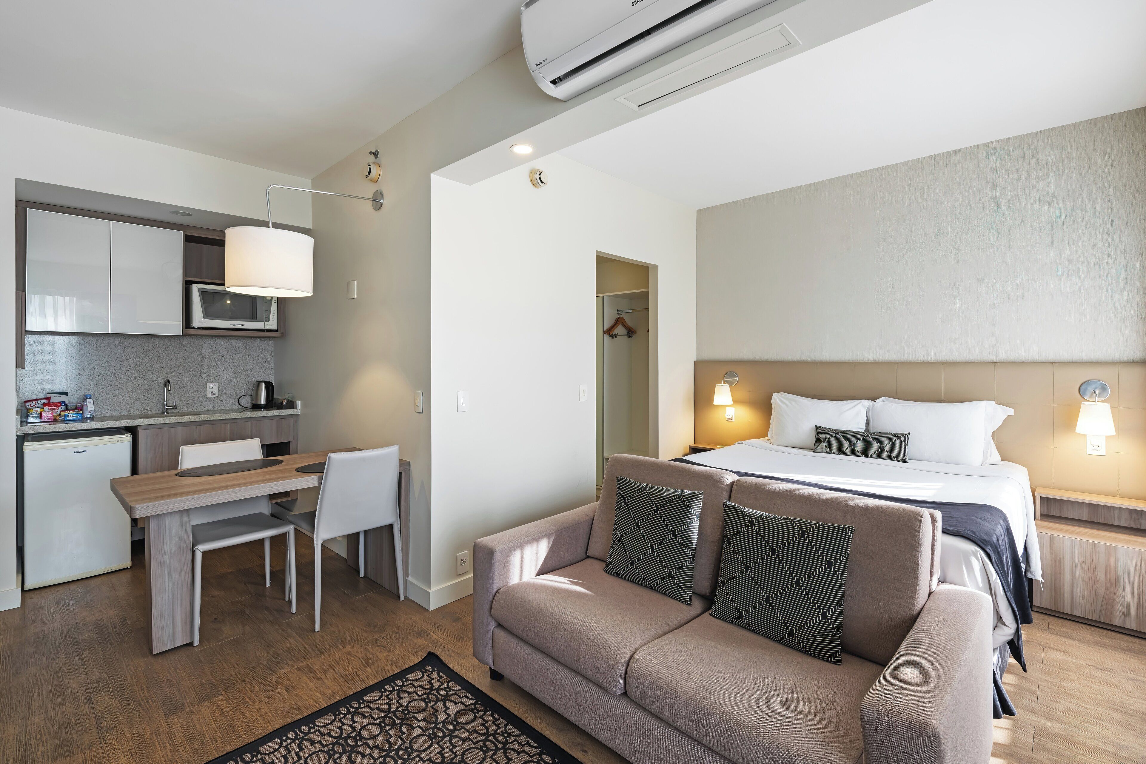 A modern hotel room with a kitchenette, a sofa, a bed, and a bedside table.