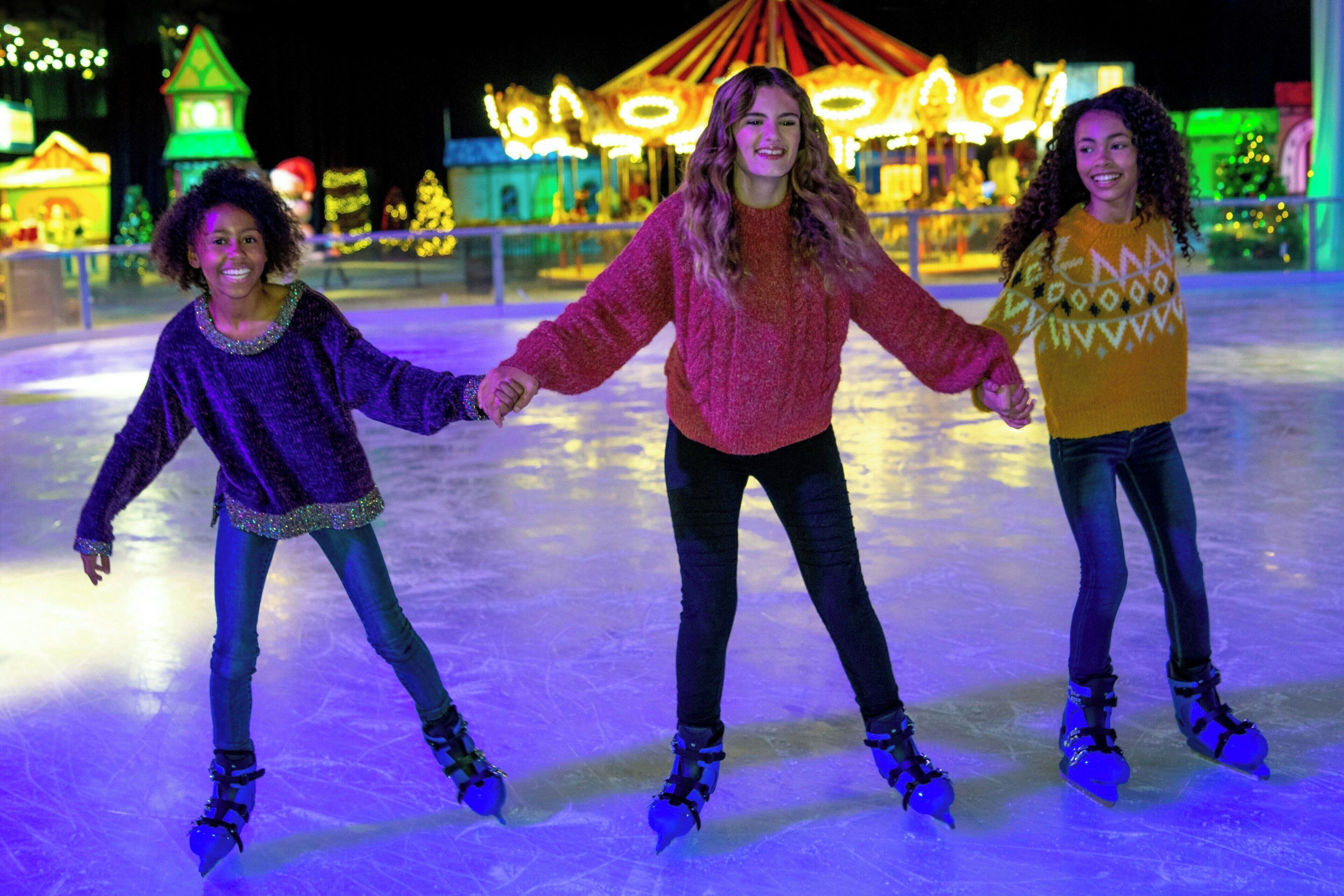 Drei Personen laufen auf einer Eislaufbahn, im Hintergrund weihnachtliche Lichter und Dekorationen.