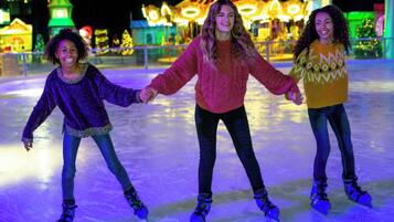 Drei Personen laufen auf einer Eislaufbahn, im Hintergrund weihnachtliche Lichter und Dekorationen.