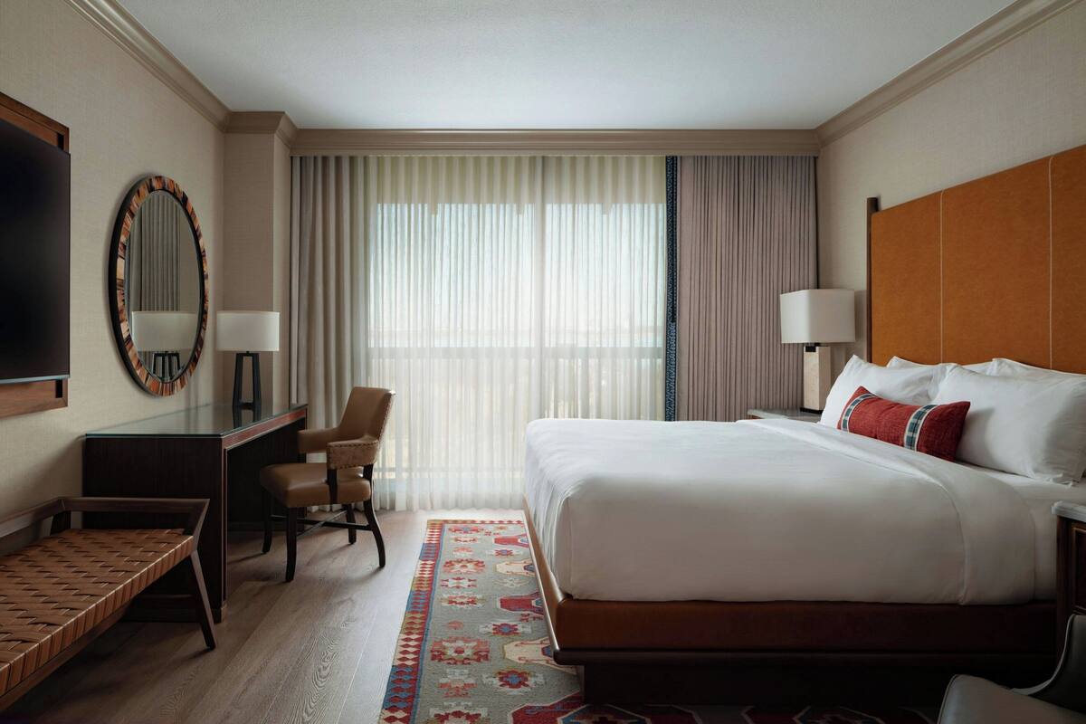 A hotel room with a bed, a desk, a chair, a TV, and a large window with curtains.