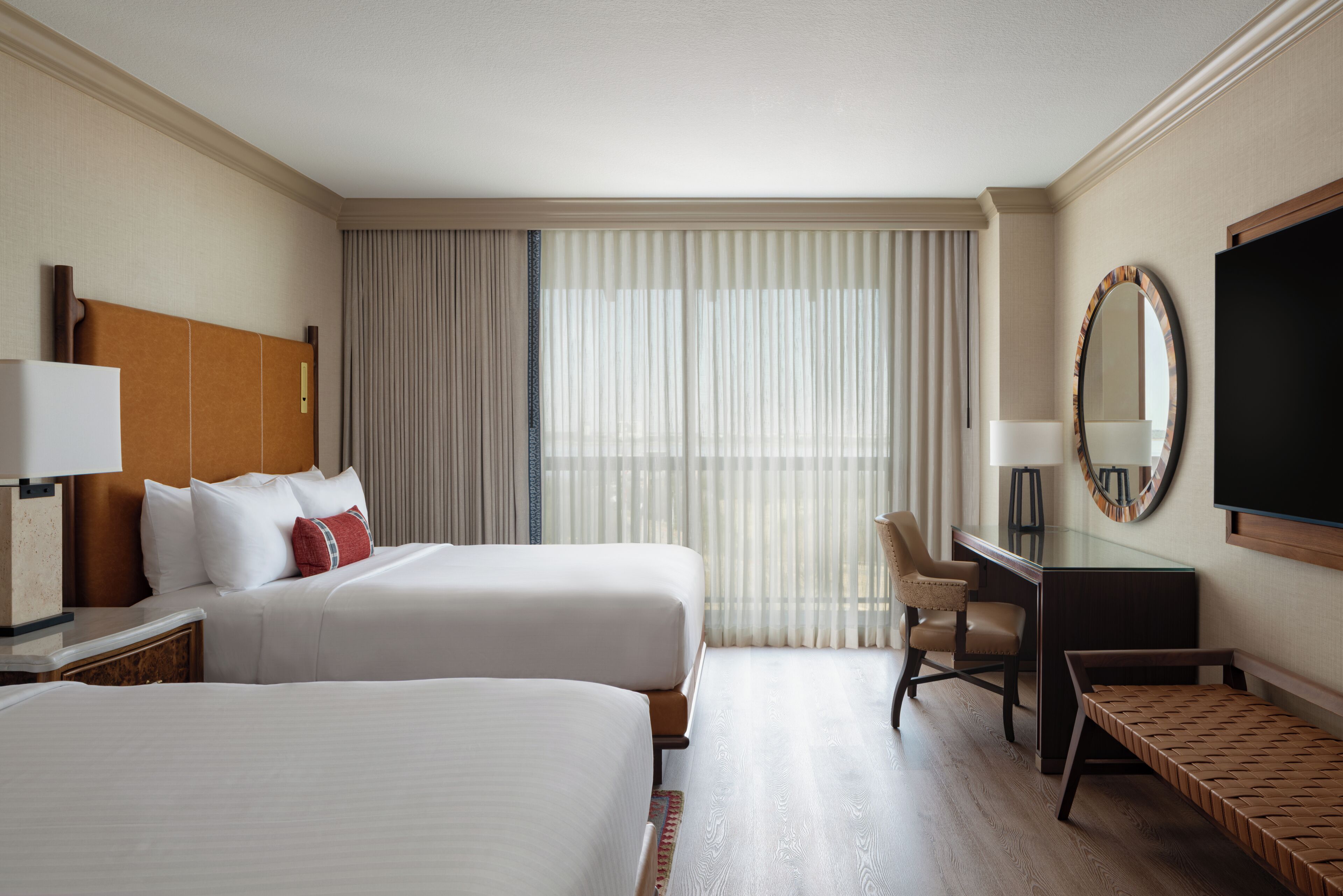 A hotel room with two beds, a desk, a chair, a TV, and a large window with curtains.