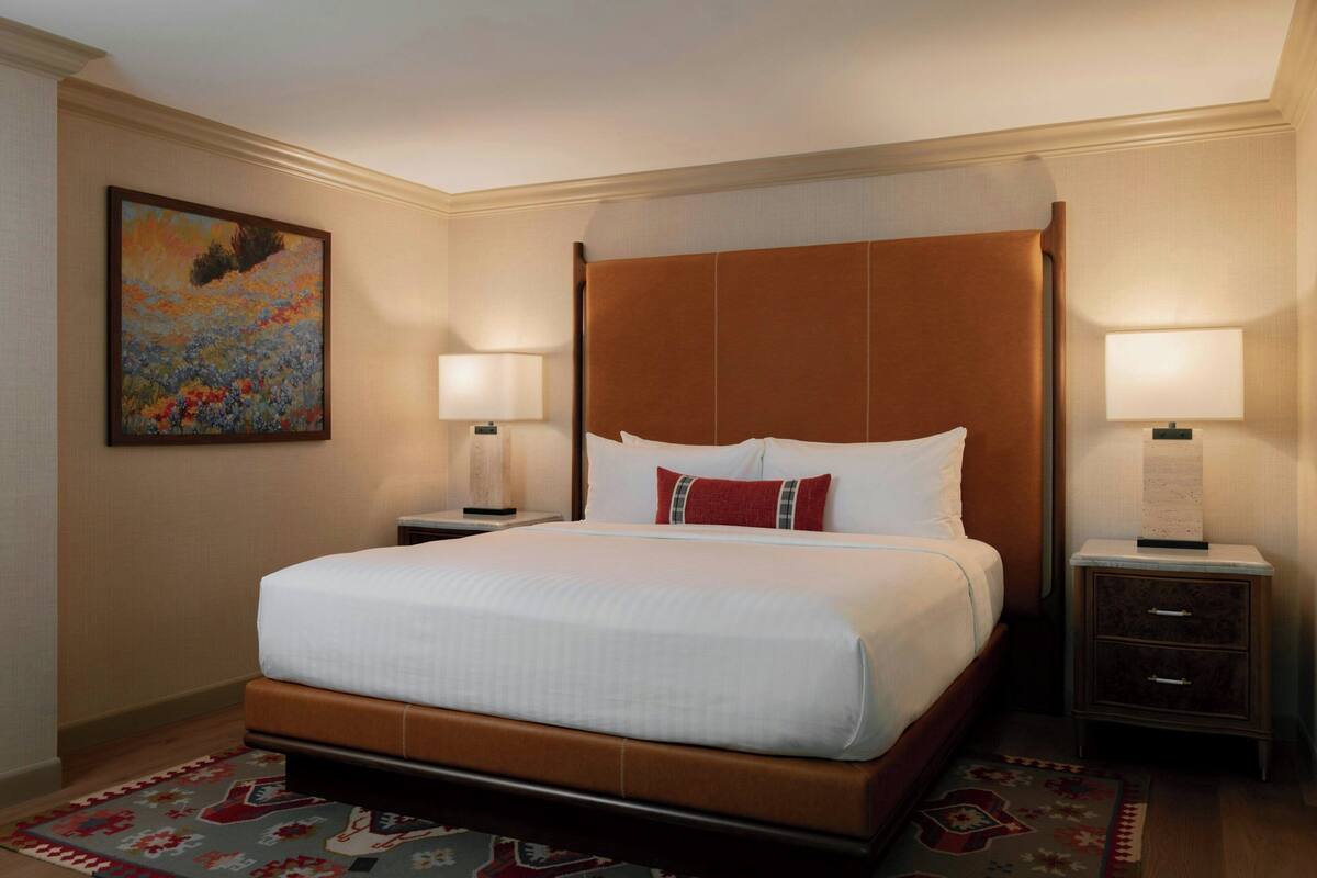 A hotel room with a large bed, two bedside tables, a painting on the wall, and a patterned rug.