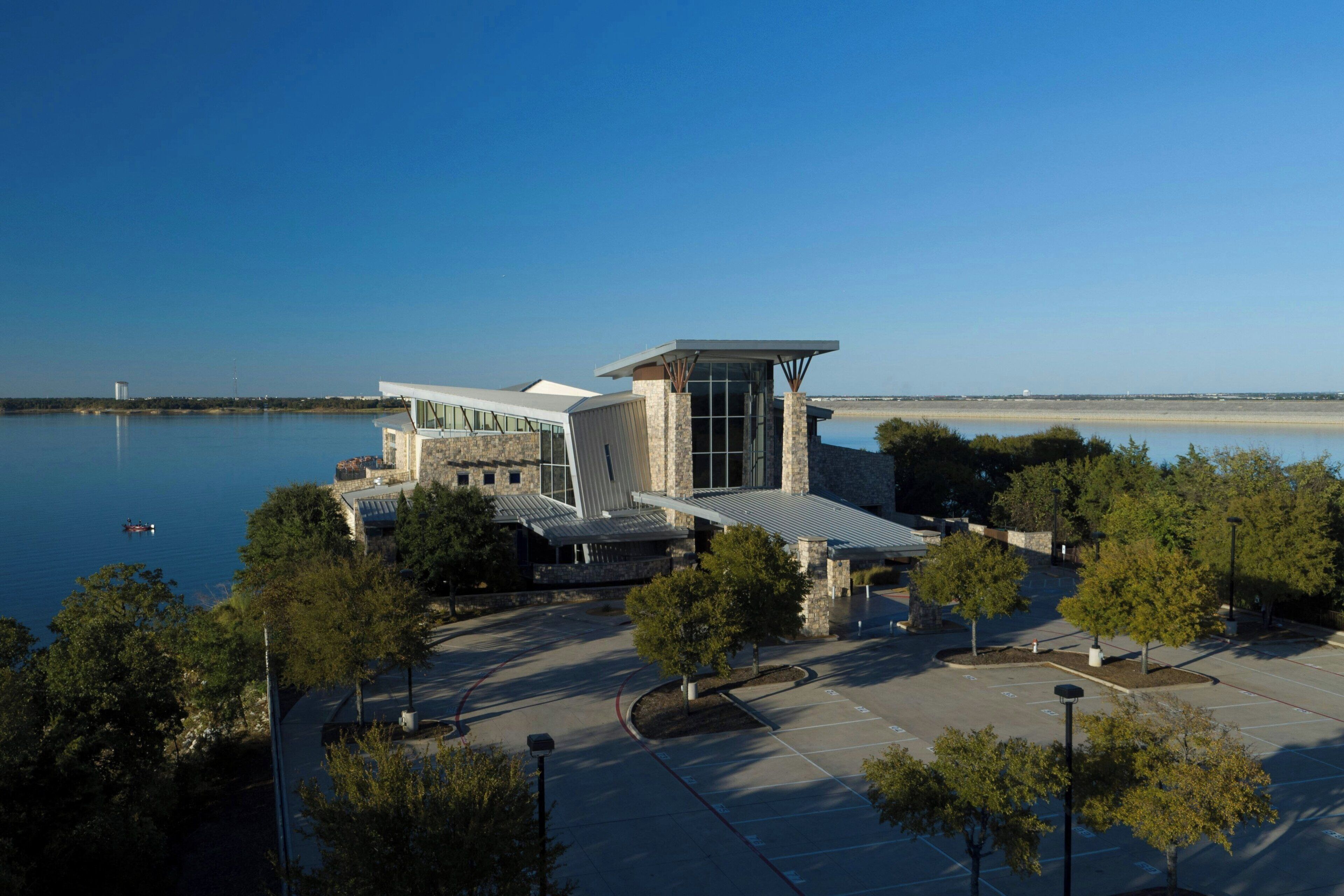 A modern building with a glass facade, surrounded by a parking lot and trees, near a calm body of water.