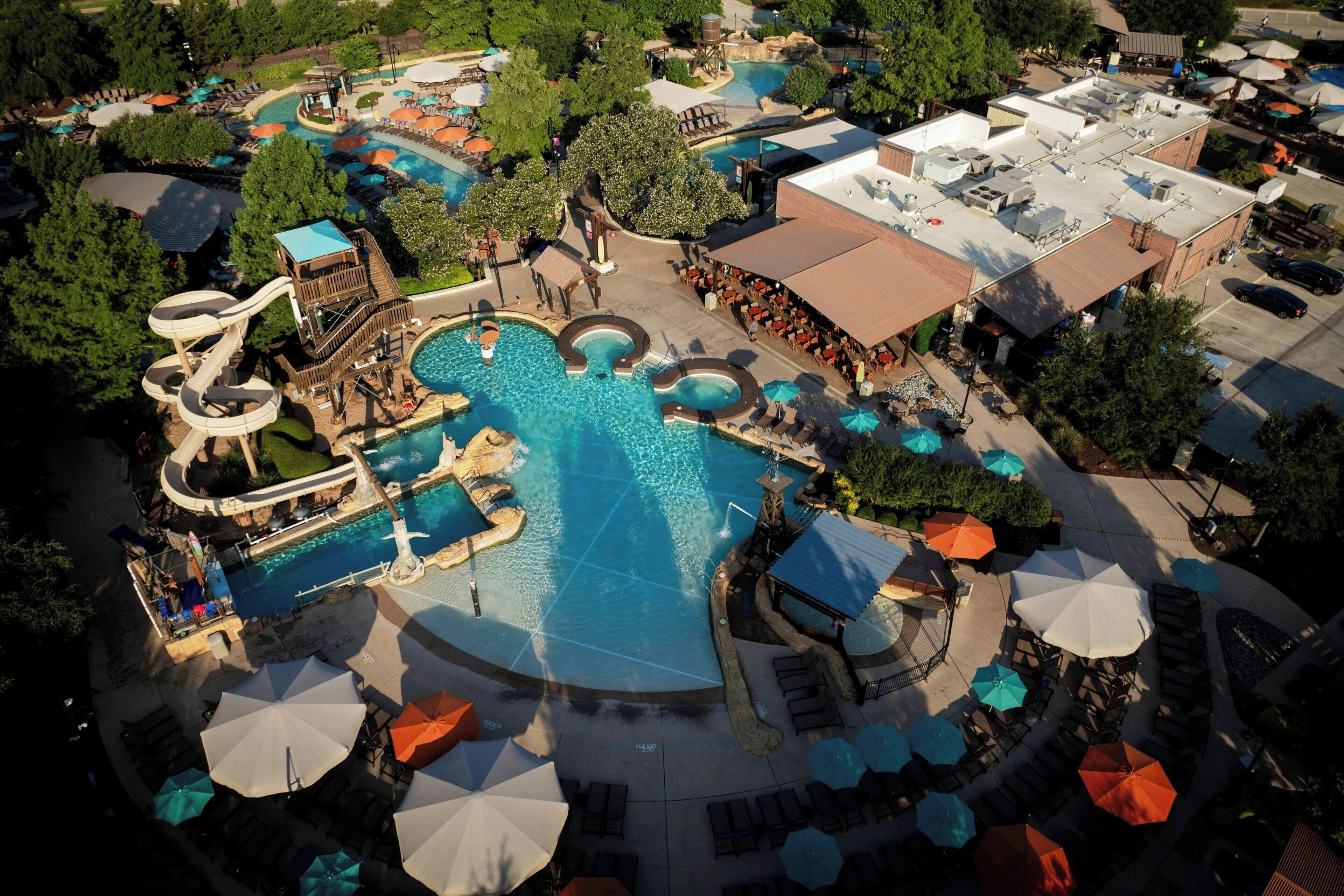 A large outdoor swimming pool with multiple slides and a water park area, surrounded by lounge chairs and umbrellas.