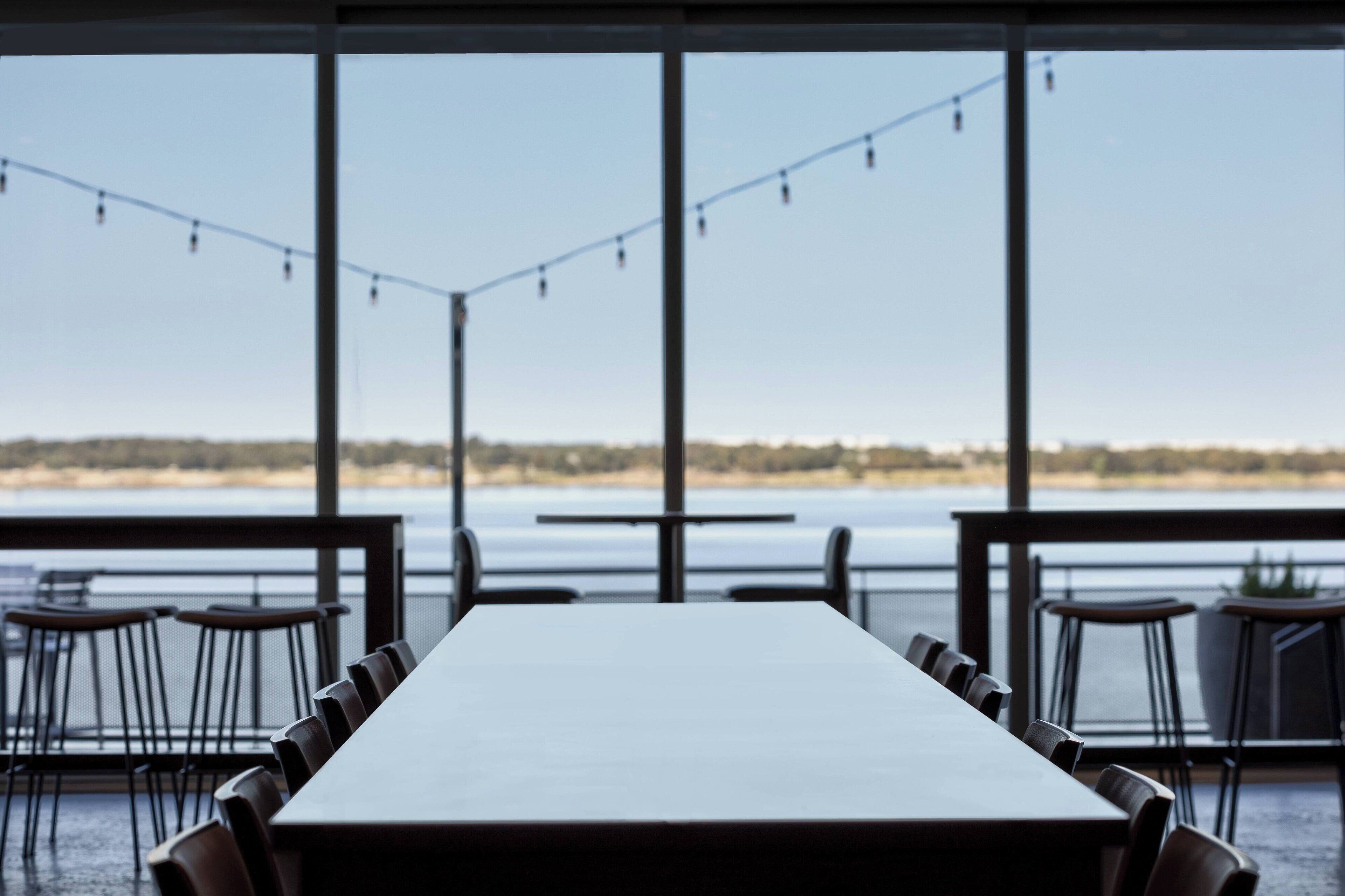 Un comedor con una mesa larga y sillas, ventanas amplias con vista a un cuerpo de agua y terreno, y luces colgantes.
