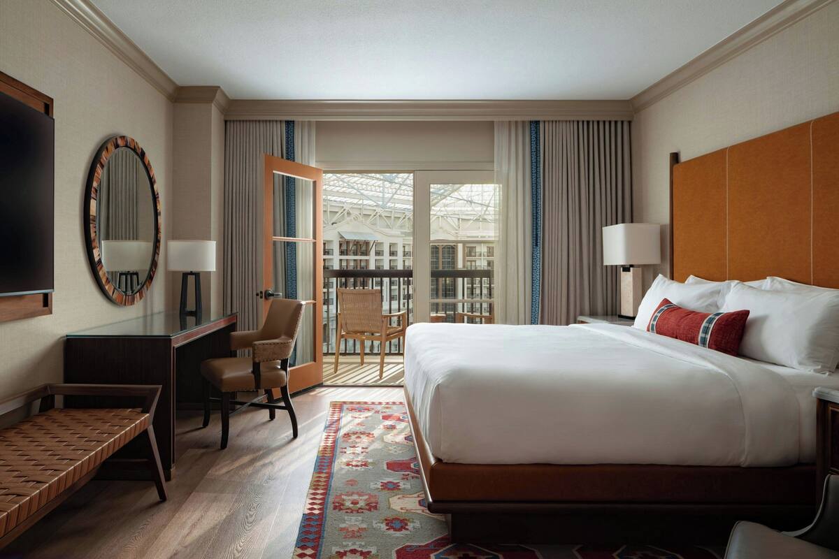 A hotel room with a large bed, a desk, a chair, a TV, and a view of a building through a glass door.