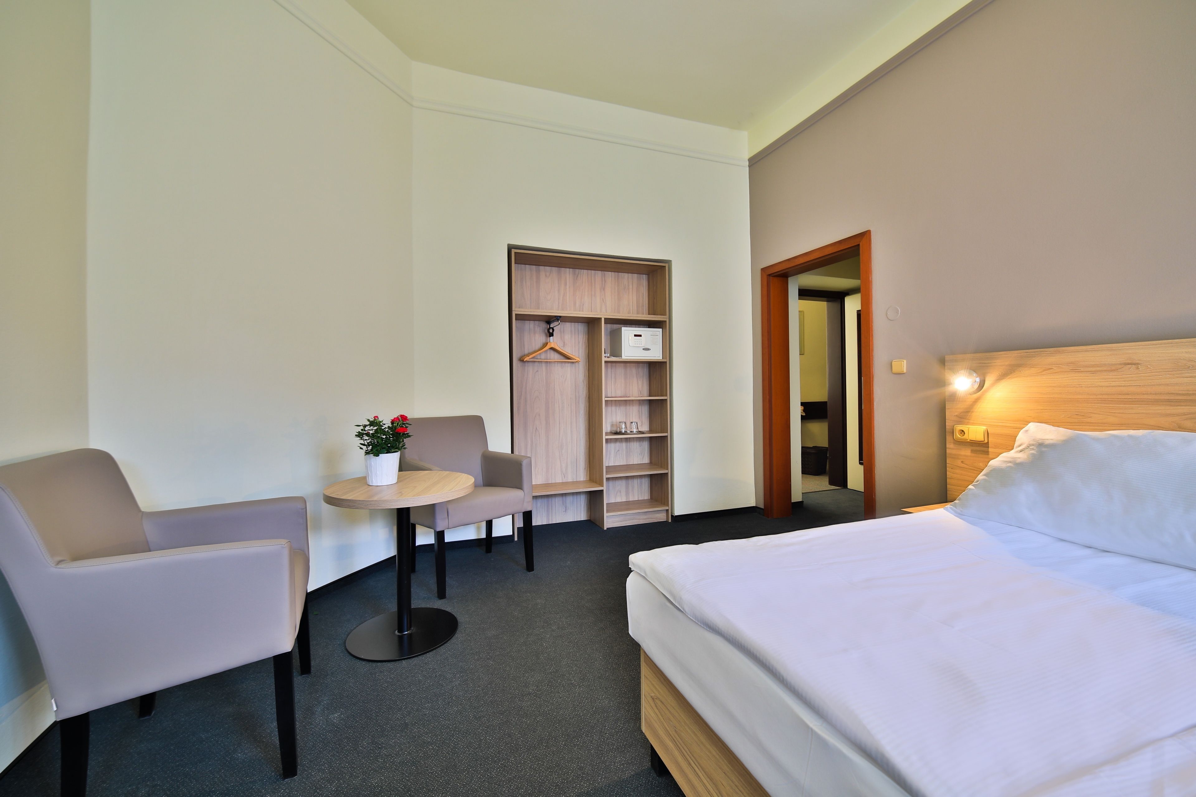 A hotel room with a bed, a desk, a chair, a small table with a plant, a wardrobe, and a mirror.