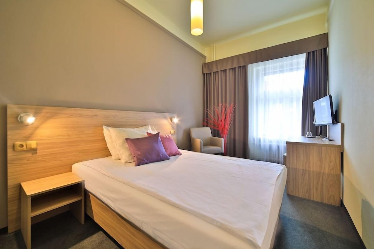 A hotel room with a large bed, bedside tables, a desk with a computer, and a window with curtains.