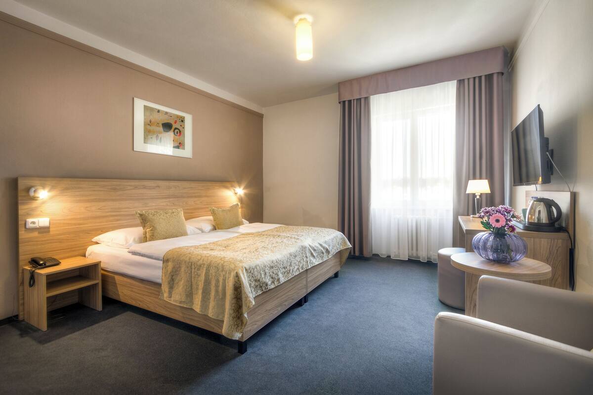 Una habitación de hotel con una cama grande, lámparas de noche, un televisor de pantalla plana, una mesita con un florero y un cuadro en la pared.