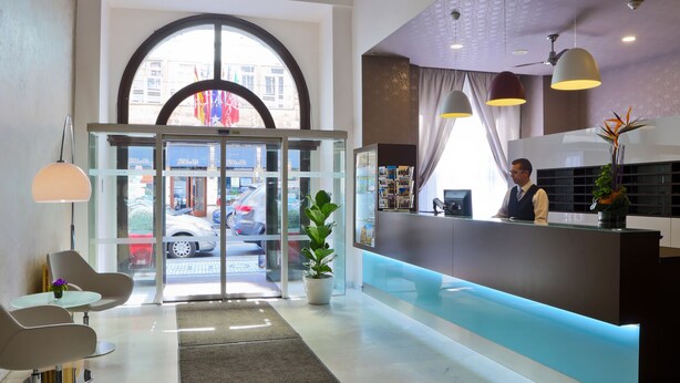 A hotel reception area with a modern design, including a reception desk, seating area, and a large window with a view of the street. - Atlantic Hotel (Prague)