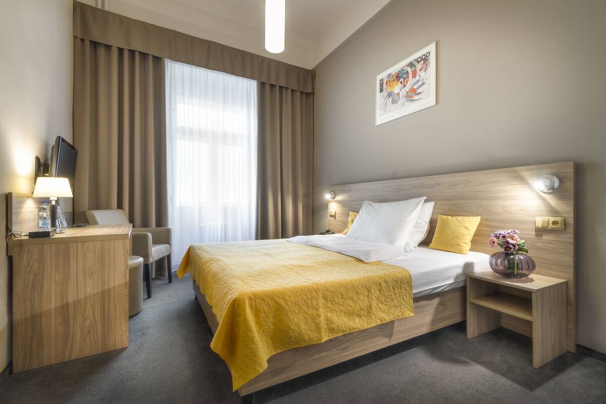 Habitación de hotel con una cama de madera, un escritorio con lámpara, una silla y una mesita de noche con un jarrón.