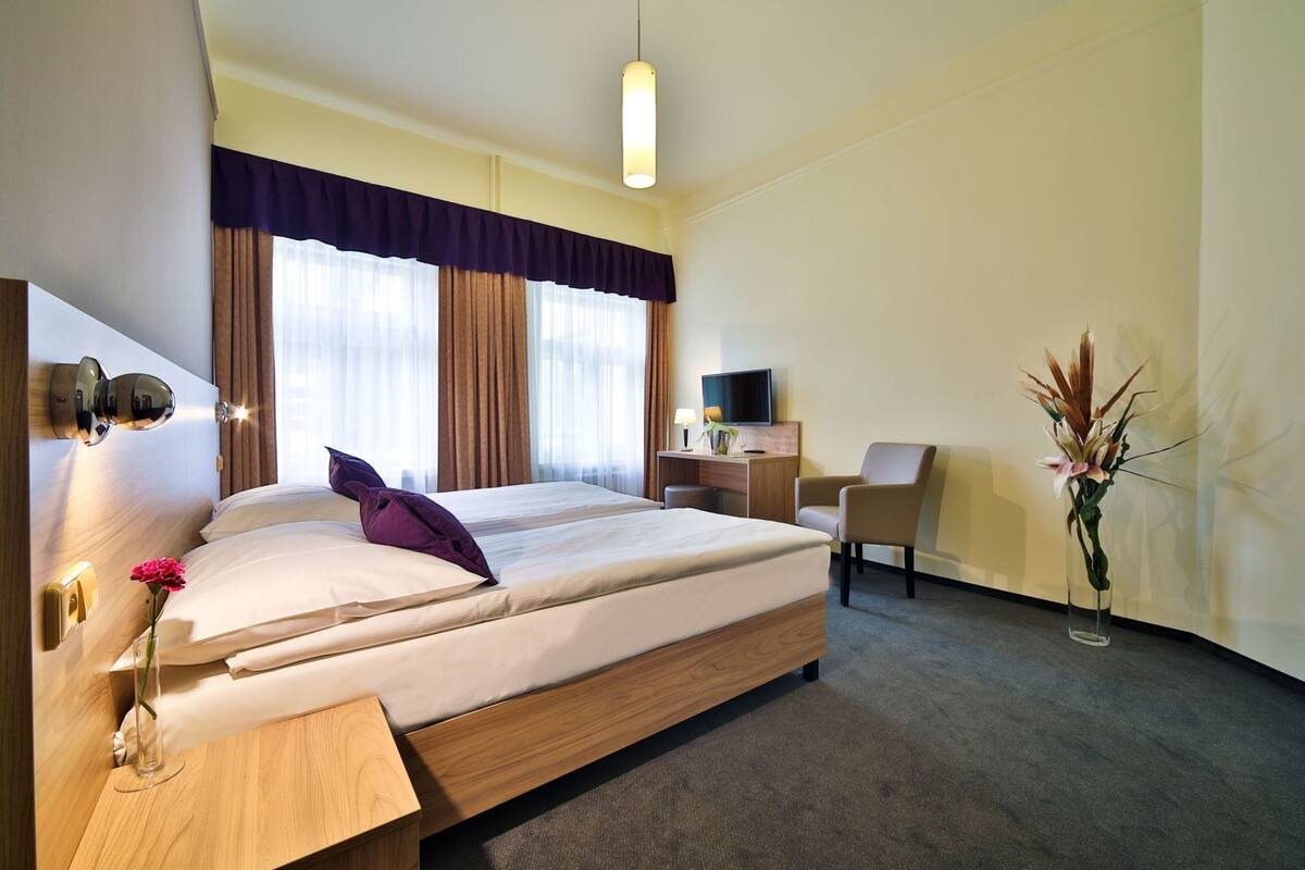 Una habitación de hotel con cama, escritorio, silla, televisor y un jarrón con flores.