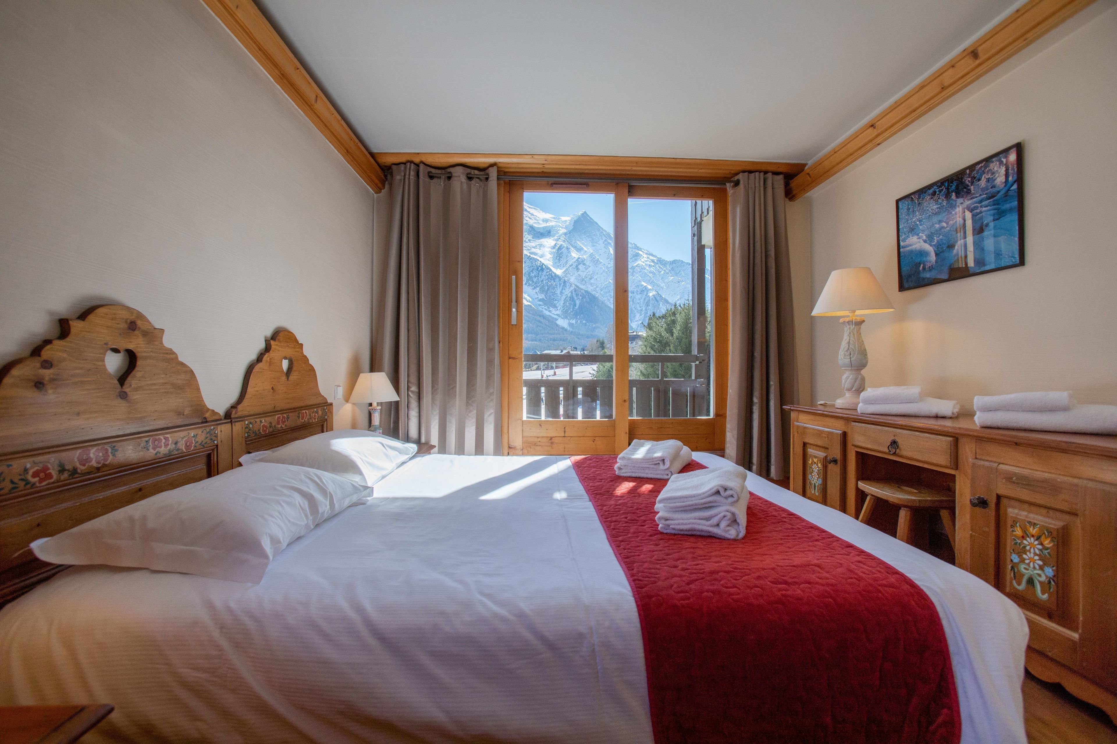 A hotel room with a bed, a wooden headboard, a nightstand, a lamp, a painting, and a view of mountains through a large window.