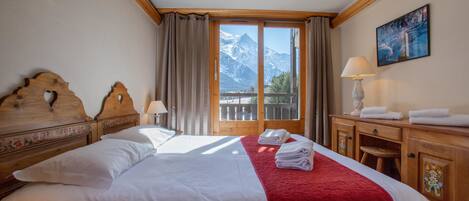A hotel room with a bed, a wooden headboard, a nightstand, a lamp, a painting, and a view of mountains through a large window.