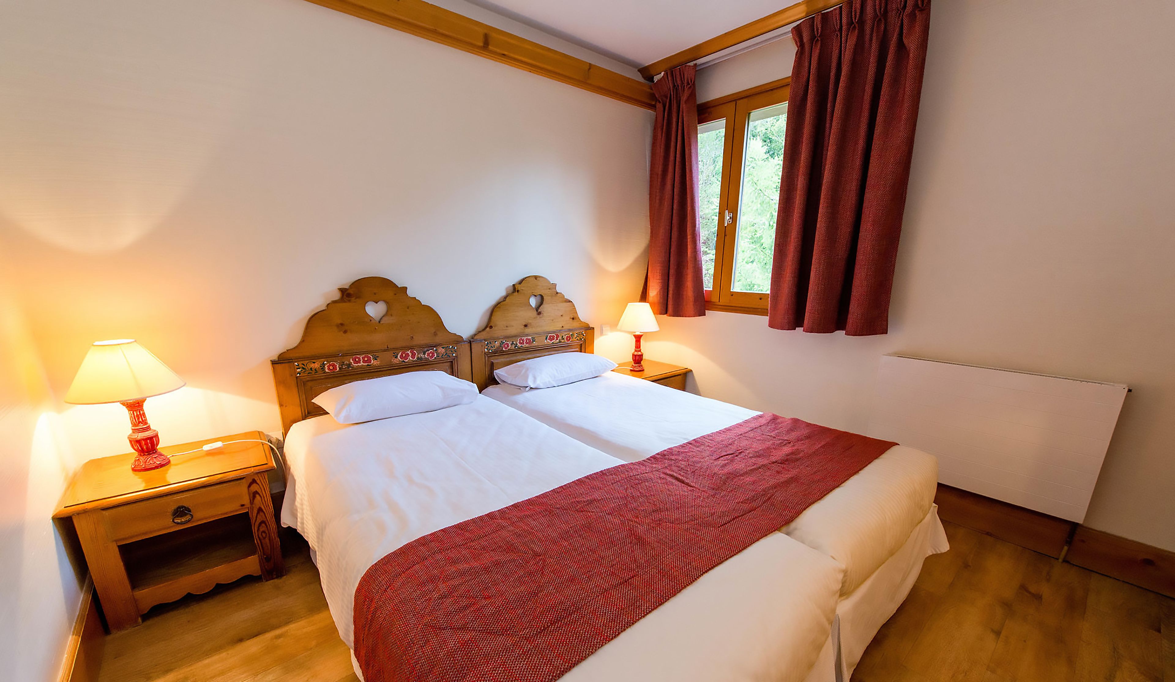 A bedroom with a wooden headboard, two bedside tables with lamps, a window with curtains, and a radiator.