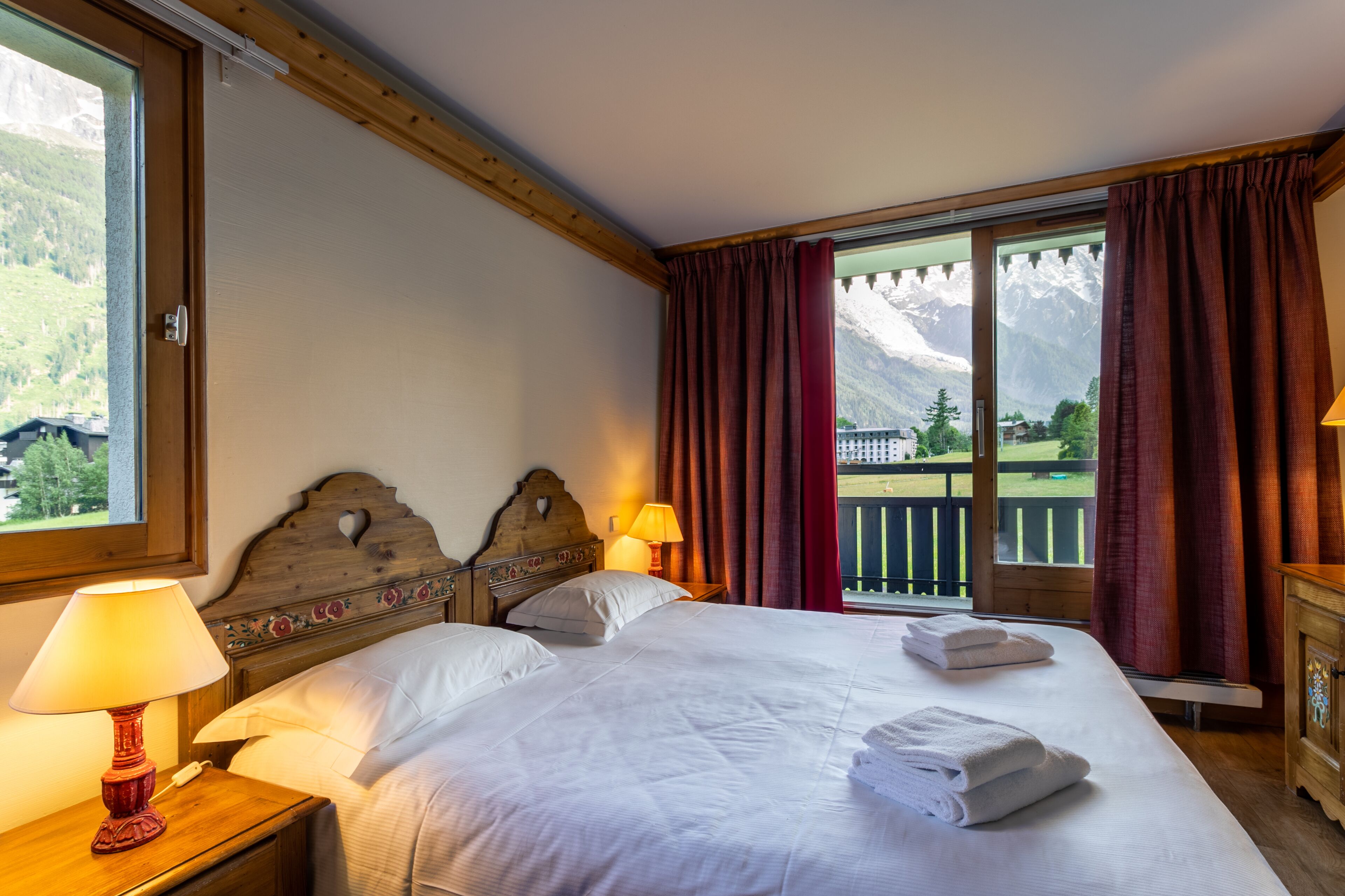 Une chambre d’hôtel avec un grand lit, deux lampes de chevet, une tête de lit en bois et une vue sur les montagnes par une fenêtre ornée de rideaux rouges.