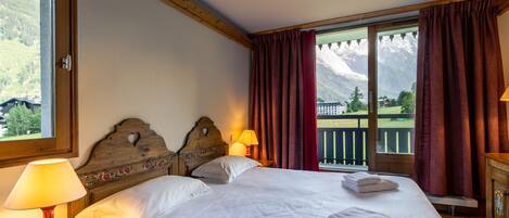 Une chambre d’hôtel avec un grand lit, deux lampes de chevet, une tête de lit en bois et une vue sur les montagnes par une fenêtre ornée de rideaux rouges.
