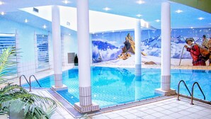 Uma piscina interior com água cristalina, rodeada por colunas brancas e um mural retratando uma paisagem montanhosa coberta de neve.