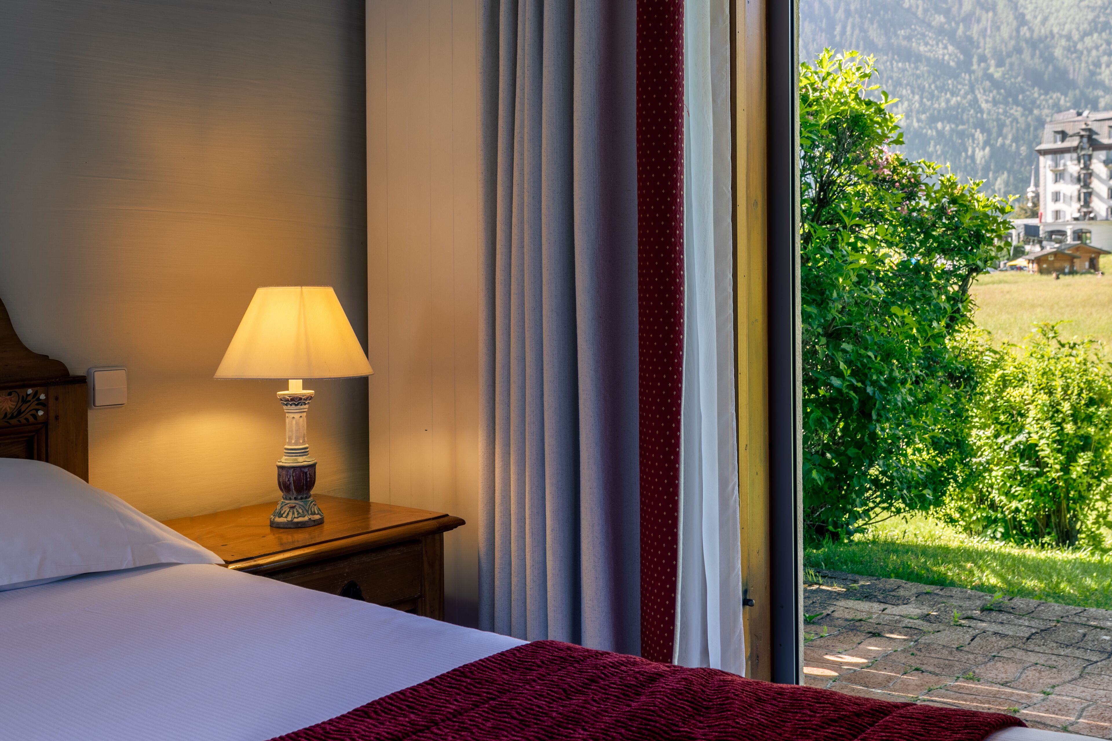 A hotel room with a bed, a bedside table with a lamp, and a view of a green landscape through a window.