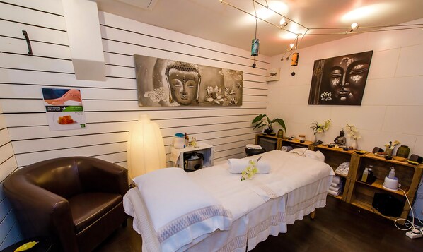 A spa room with a massage table, a massage chair, a shelf with spa products, and two wall-mounted paintings. - Les Balcons du Savoy (Chamonix-Mont-Blanc)