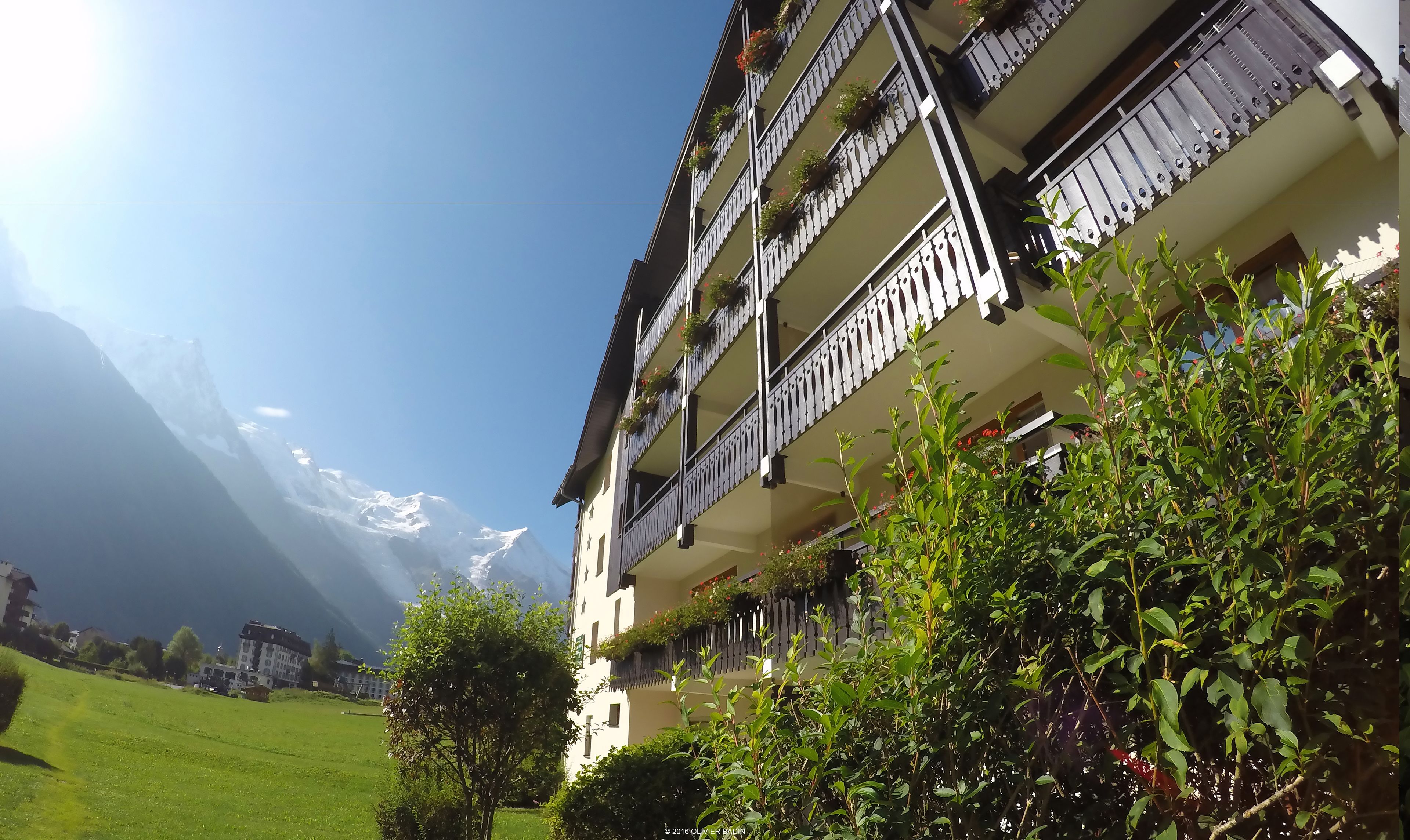 Un edificio de varios pisos con balcones, rodeado de vegetación y montañas al fondo.