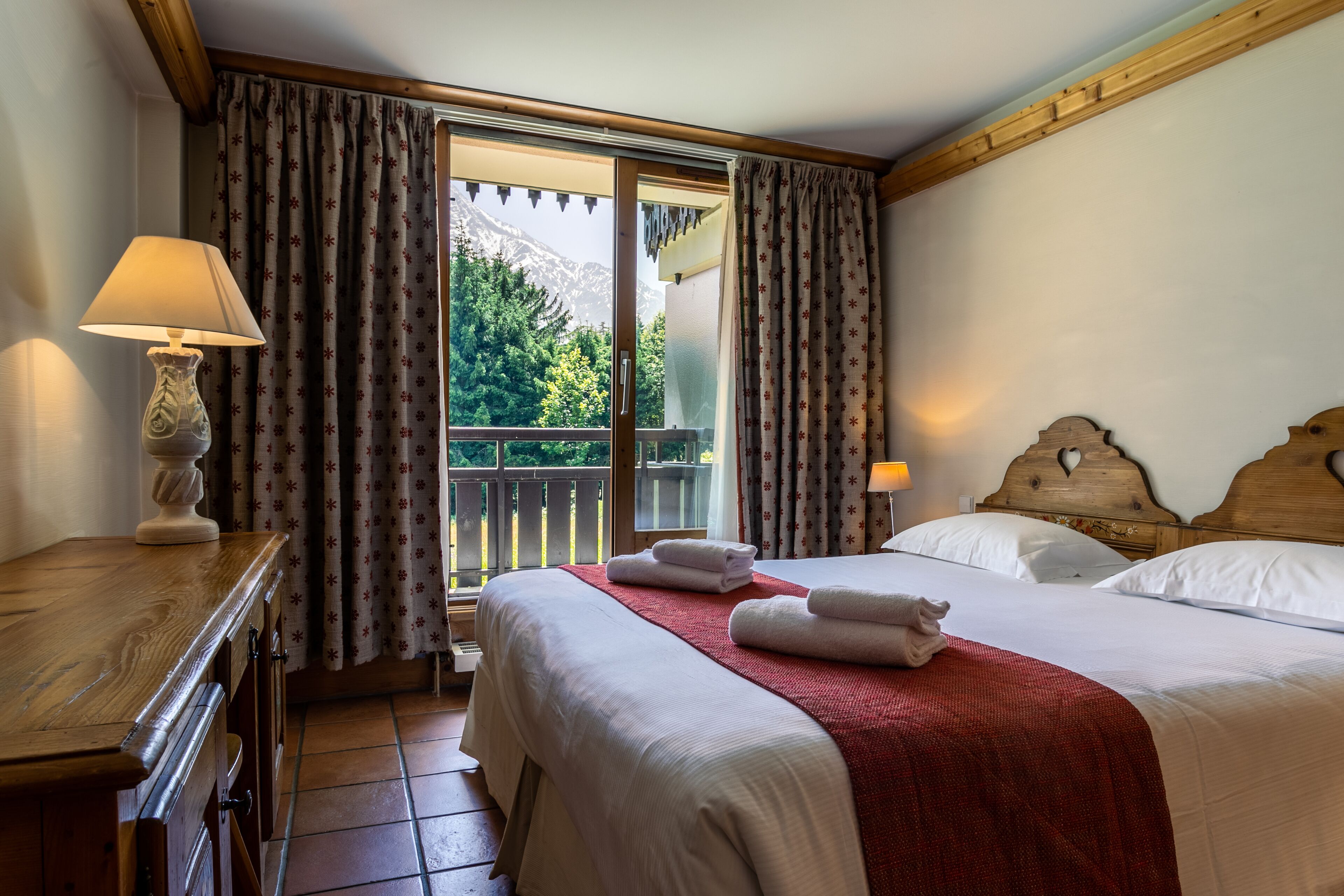 A hotel room with a bed, a wooden nightstand, a lamp, and a view of a balcony with curtains.
