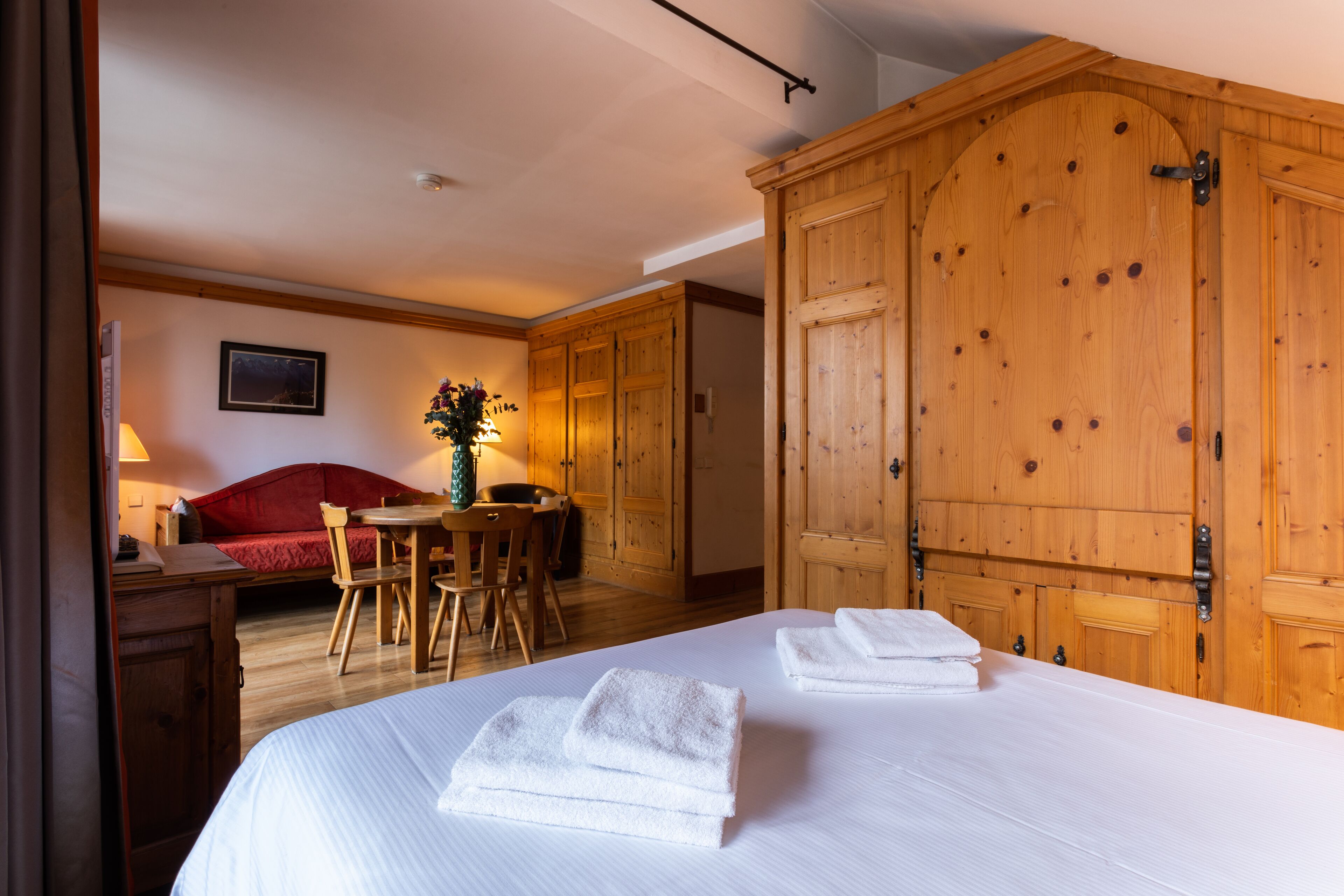 Habitación de hotel con una cama grande, armario de madera, sofá rojo, mesa de comedor con sillas y una lámpara.
