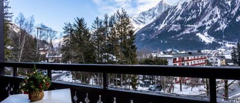 A balcony with a view of snow-covered mountains, a town in the background, and a table with a potted plant.