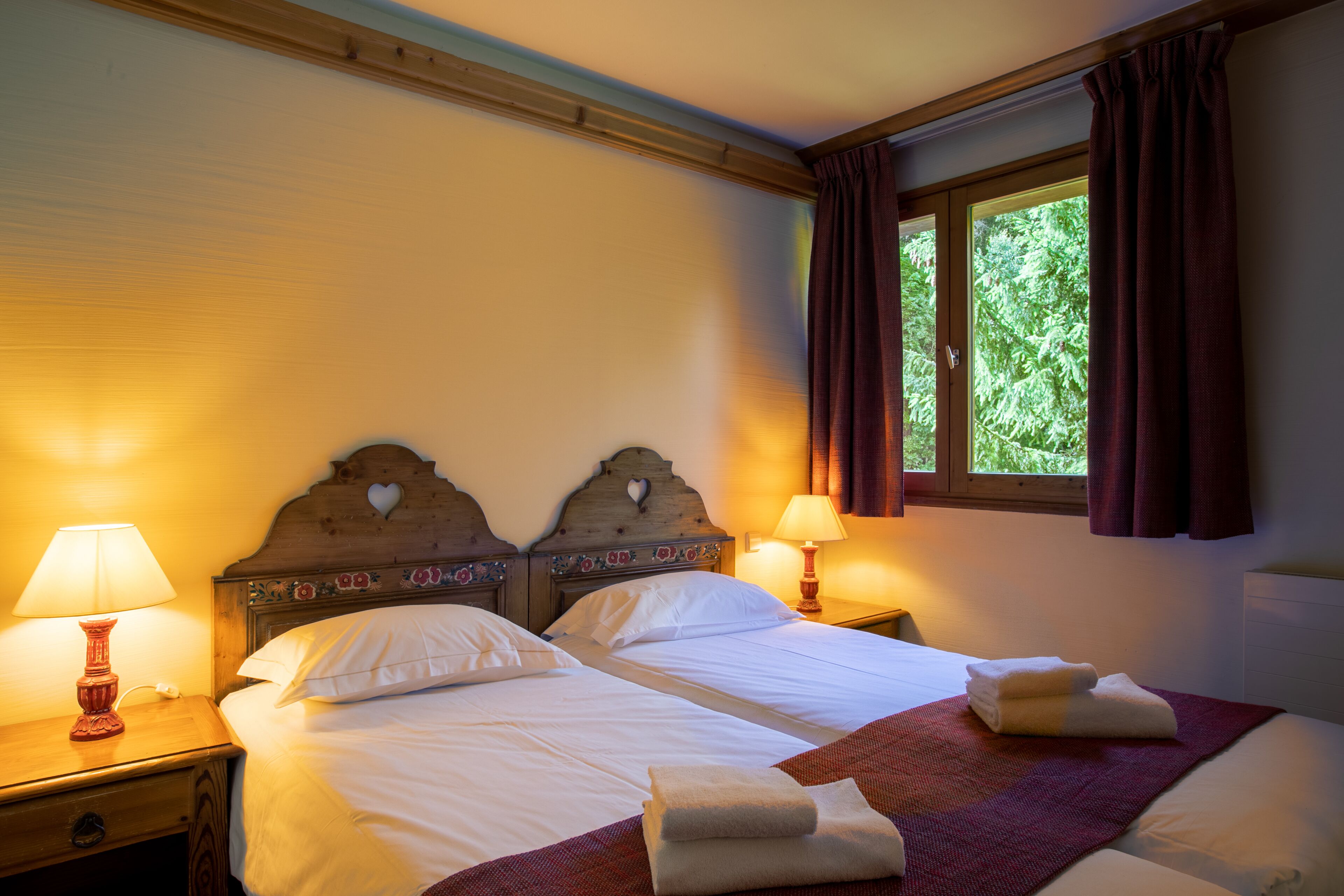 Una cama doble con ropa de cama blanca, dos mesitas de noche con lámparas, un ventanal con cortinas y un cabecero de madera con detalles florales tallados.