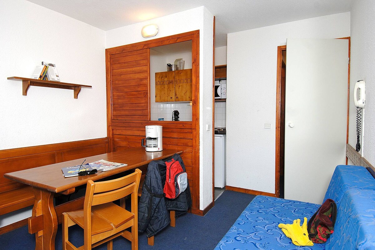 A compact room with a wooden table, a single chair, a bed with a blue cover, a shelf with books, and a kitchenette area.
