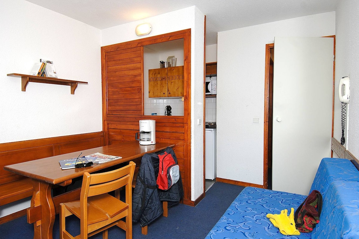 Une petite pièce comprenant une table en bois, une seule chaise, un lit recouvert d’une couverture bleue, une étagère remplie de livres et un coin cuisine.