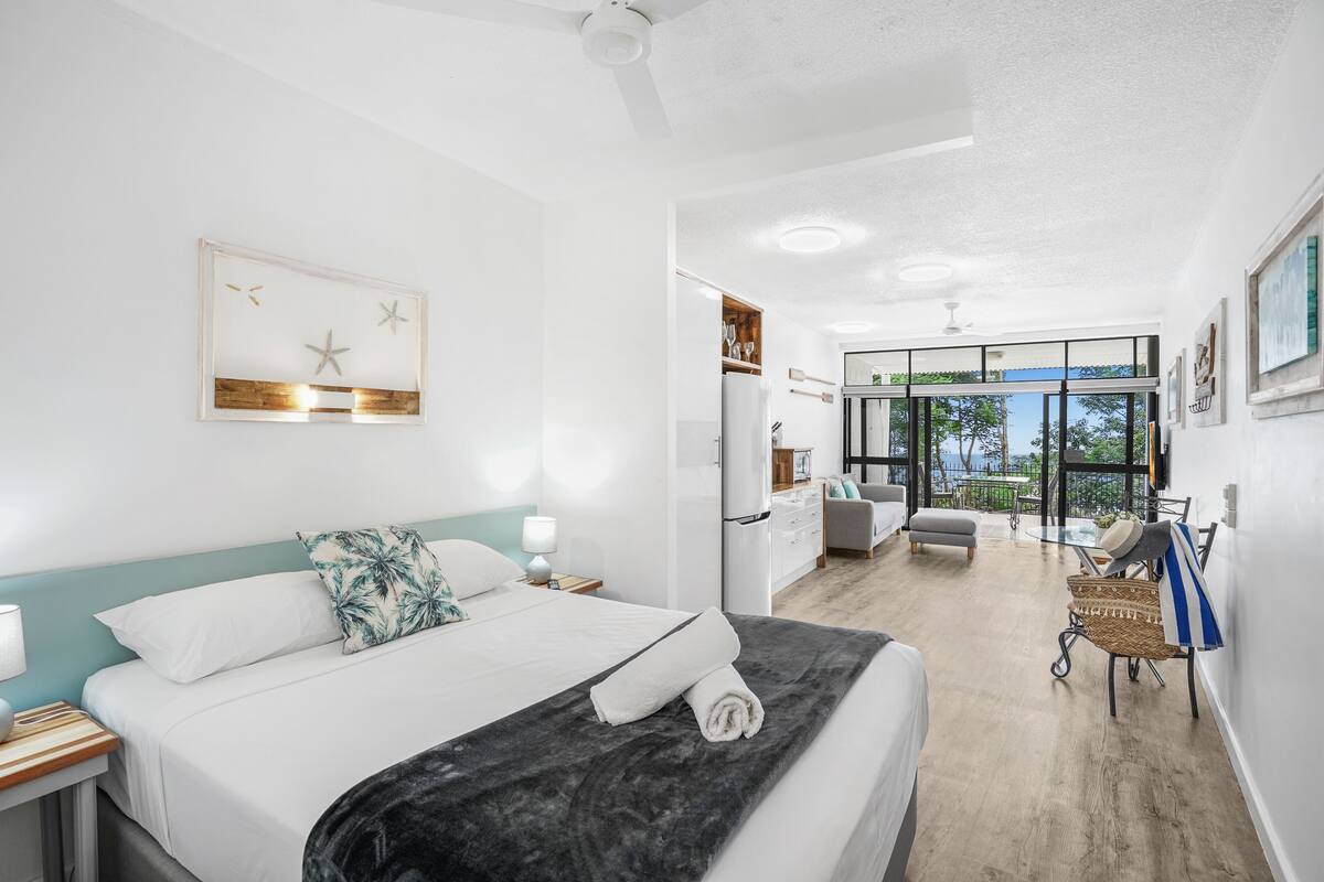 A modern hotel room with a large bed, a desk, a chair, and a balcony with a view.