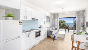 Una cocina moderna con armarios blancos, un lavamanos y una estufa. Hay un comedor con mesa y sillas, y un balcón con vista al océano.