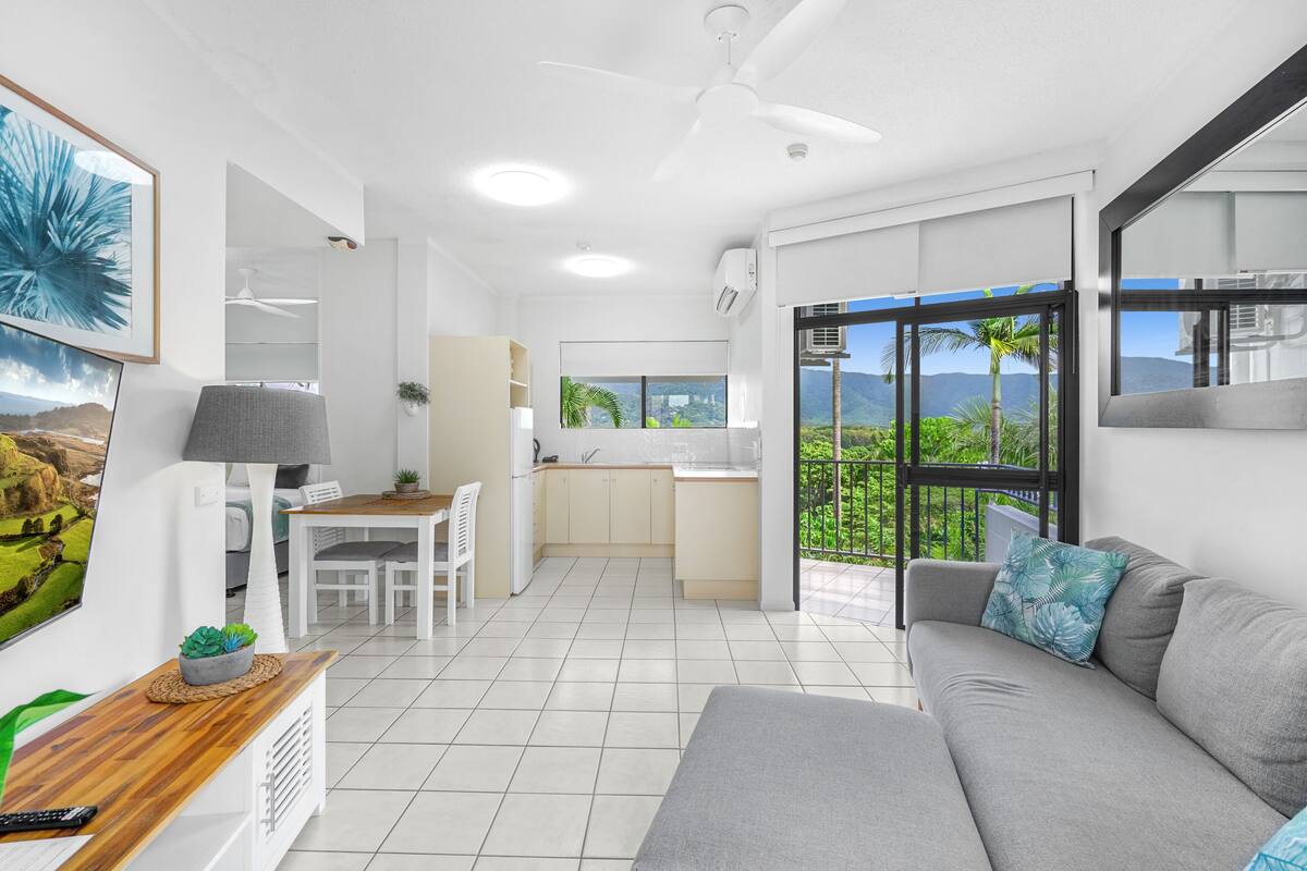 A modern living room with a grey sofa, a dining area, and a kitchen with a view of greenery.