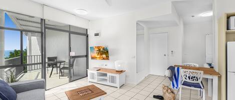 A modern living room with a sofa, a wooden table, a TV, and a balcony with a view.