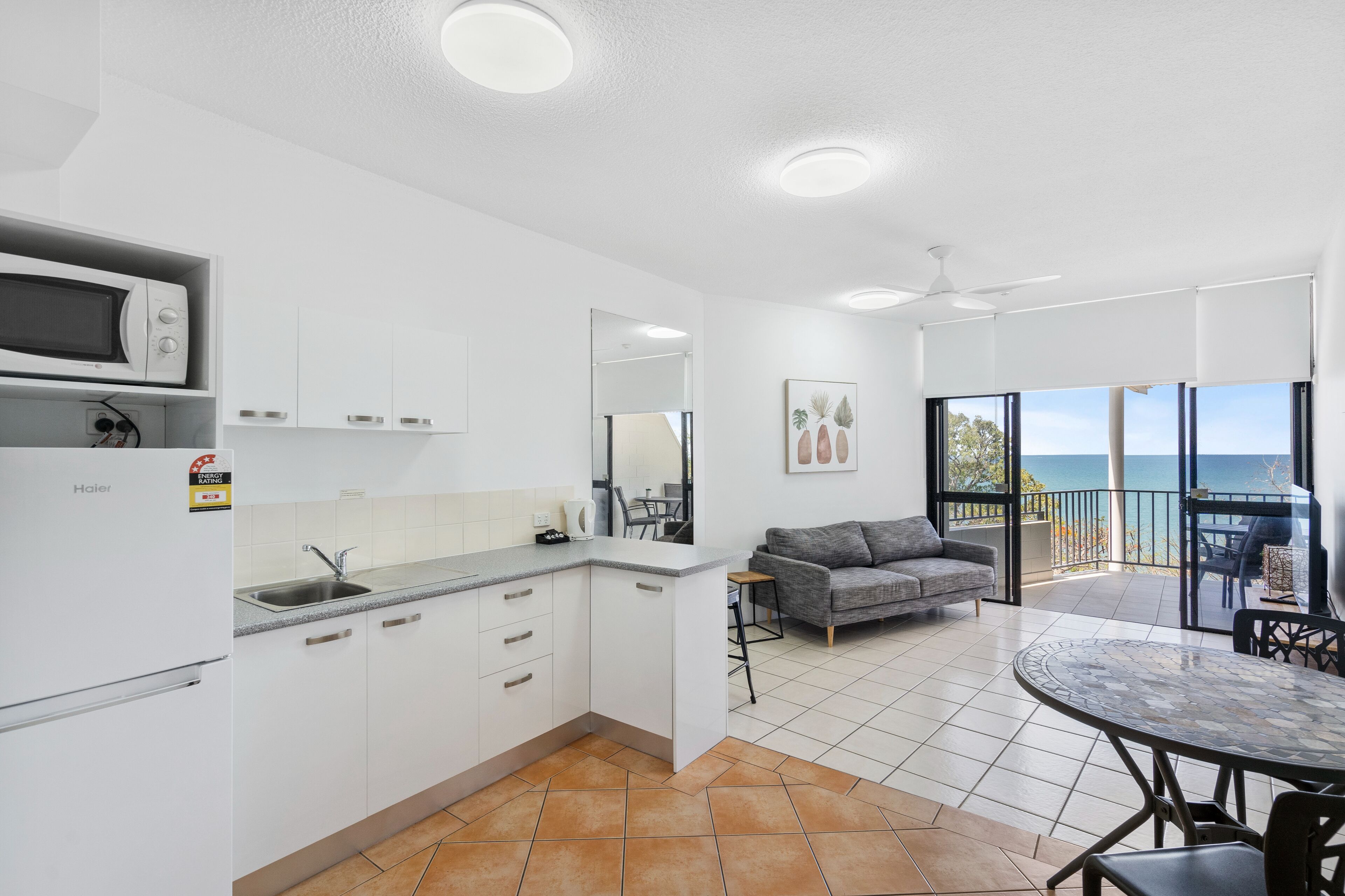 Una cocina moderna con armarios blancos, un microondas y un fregadero. En la zona de estar, hay un sofá gris y una mesa de comedor con sillas. La habitación tiene un ventanal con vistas al mar.