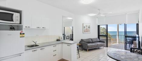Una cocina moderna con armarios blancos, un microondas y un fregadero. En la zona de estar, hay un sofá gris y una mesa de comedor con sillas. La habitación tiene un ventanal con vistas al mar.