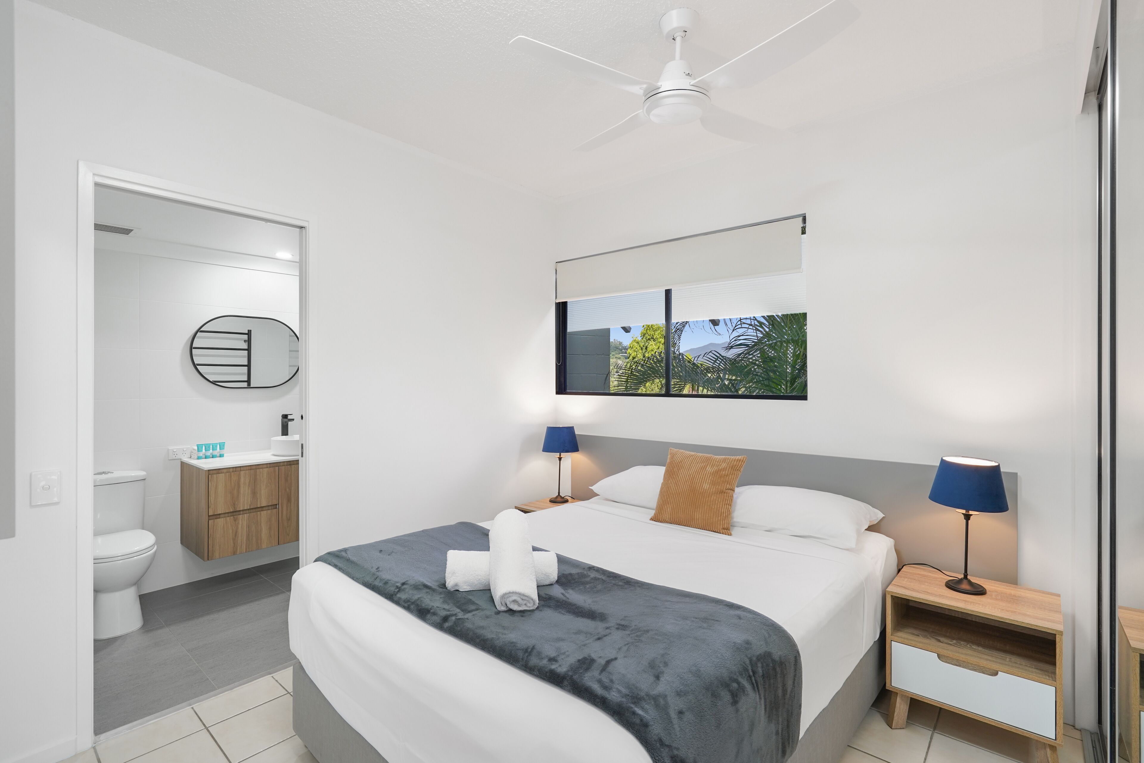 Une chambre d’hôtel moderne dotée d’un grand lit, d’un ventilateur de plafond, d’une salle de bain avec des toilettes et un lavabo, et offrant une vue sur la verdure par la fenêtre.