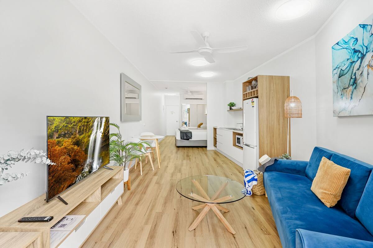A modern living room with a flat-screen TV, a glass coffee table, a blue sofa, wooden flooring, and a ceiling fan.