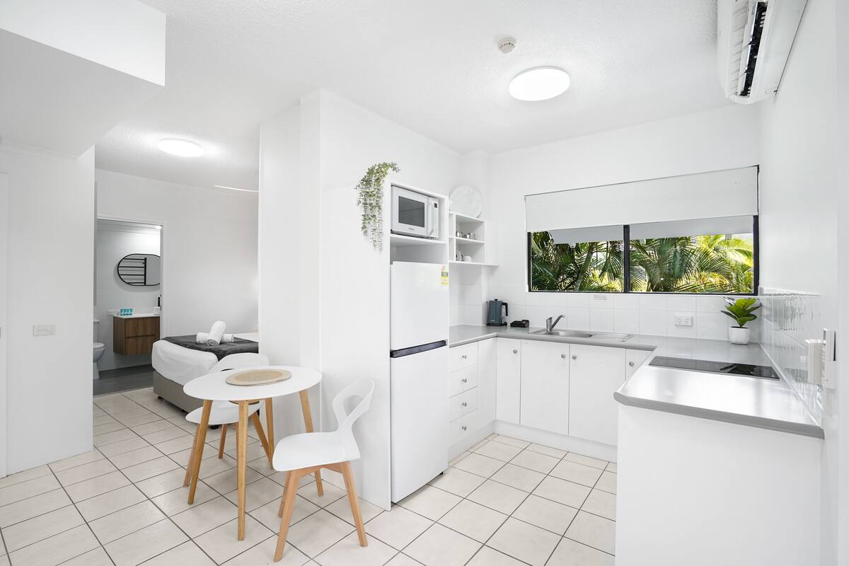 A modern kitchen with white cabinetry, a dining area with a small table and chairs, and a view of greenery through a window.