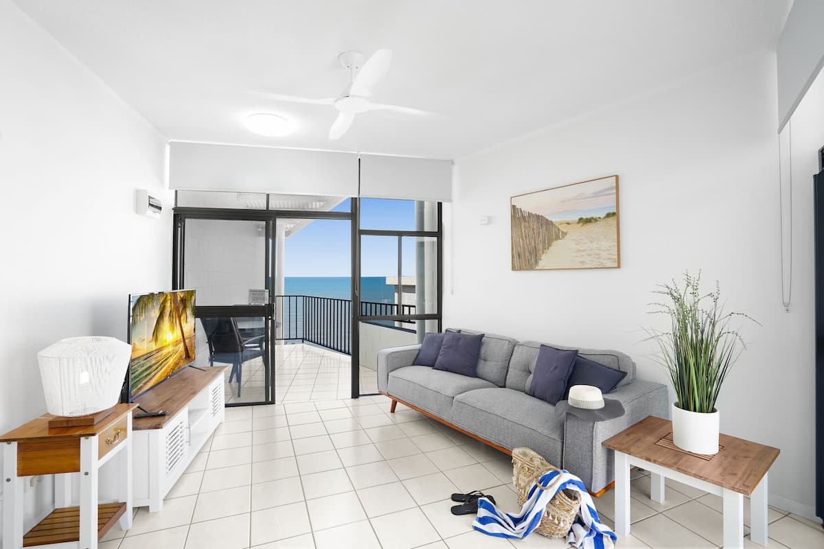 A modern living room with a grey sofa, a wooden coffee table, and a TV on a white stand. There is a balcony with a glass door leading to a terrace with a view of the sea.