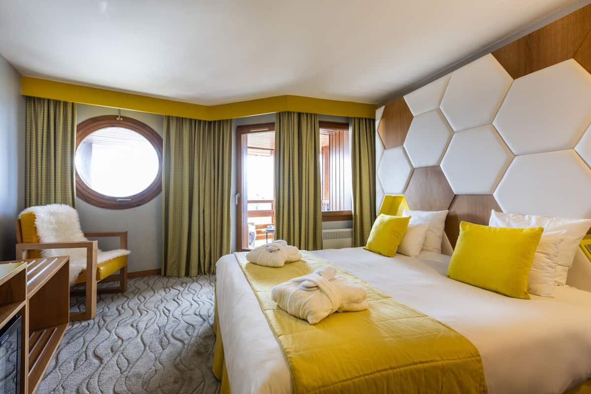 Ein Hotelzimmer mit einem großen Bett, einem Schreibtisch und einem Stuhl. Es gibt ein Fenster mit Vorhängen, einen Blick nach draußen und ein auffälliges, sechseckiges Kopfteil.