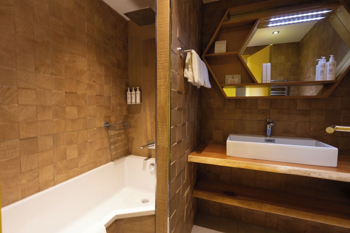 A modern bathroom with a wooden wall, a white bathtub, a sink with a mirror above it, and a shelf with toiletries.