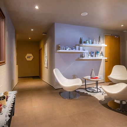 A modern hotel lobby with a reception desk, white armchairs, a coffee table, a glass display case, and a wall-mounted shelf with toiletries.