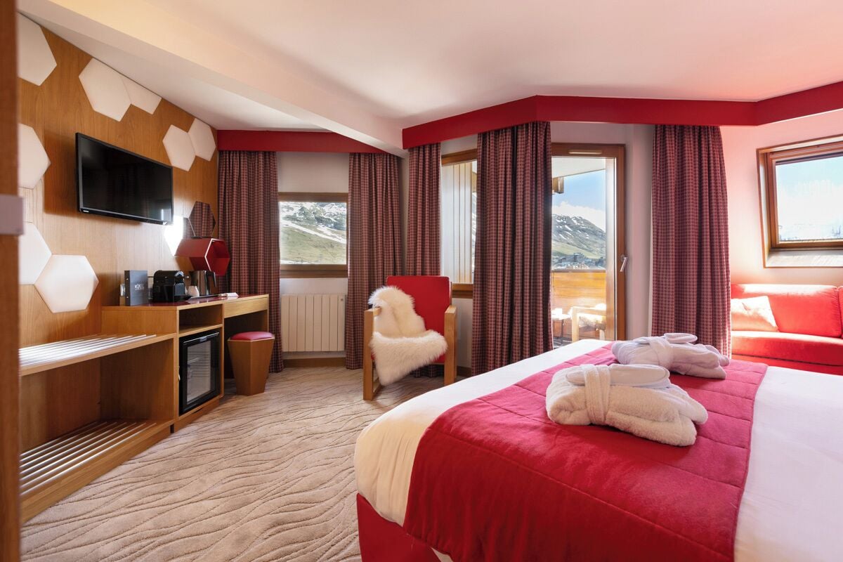 Habitación de hotel con una cama grande, un escritorio con computadora, una silla, televisor y ventana con cortinas.