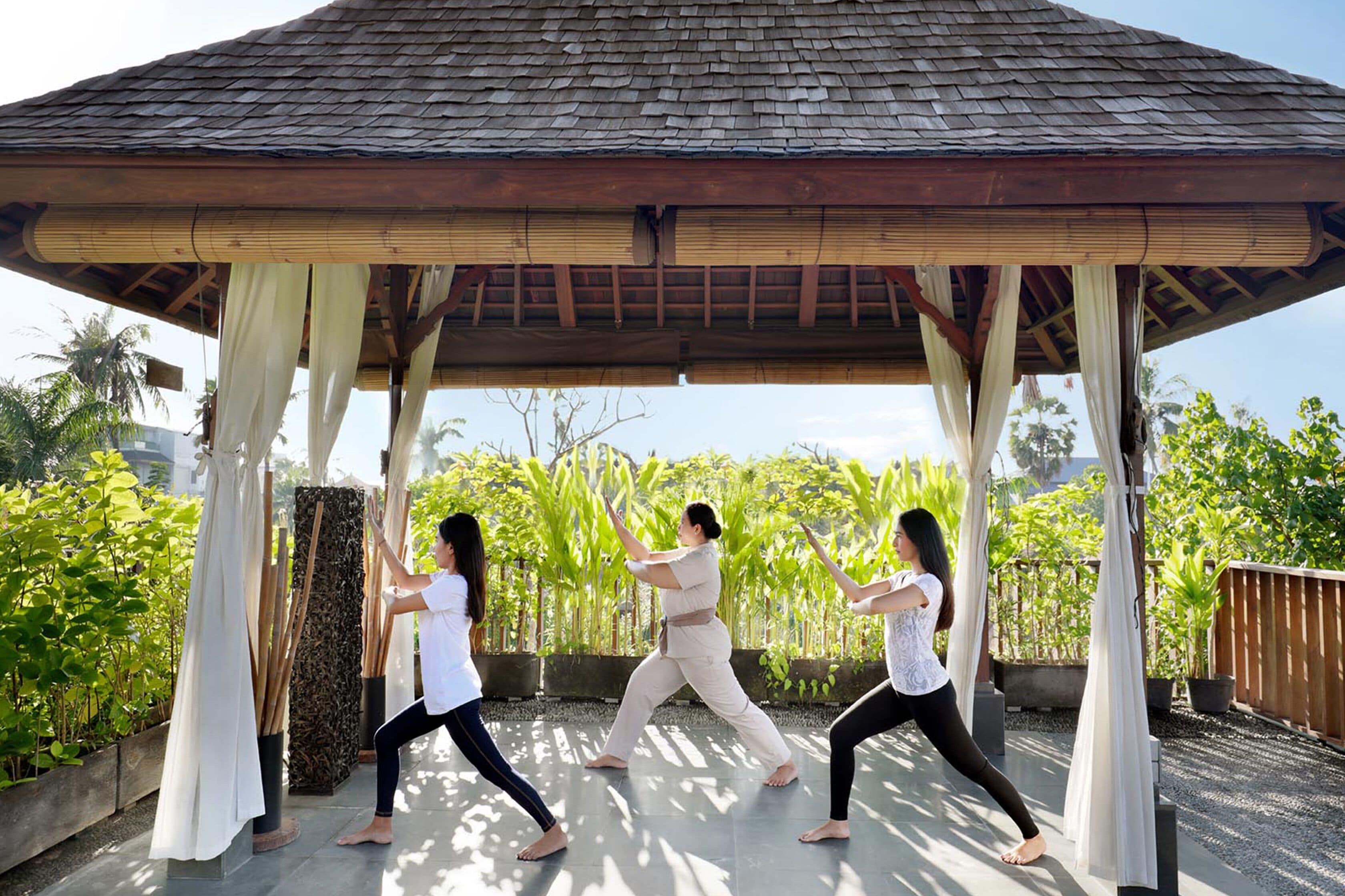Three people practicing yoga under a wooden gazebo with a tiled floor, surrounded by greenery.