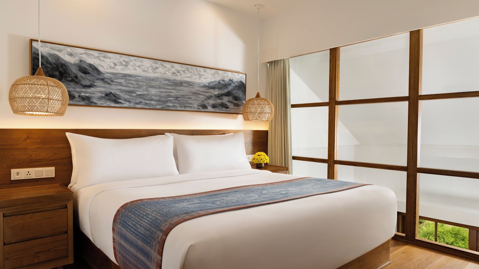A hotel room with a large bed, wooden headboard, a painting above the bed, a nightstand with a lamp, and a large window with a view of greenery.